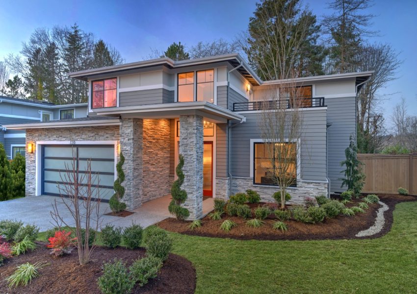 Modern contemporary house exterior with luxury details, landscaping, stone, wood, glass, lots of large windows with evening lights and red front door.